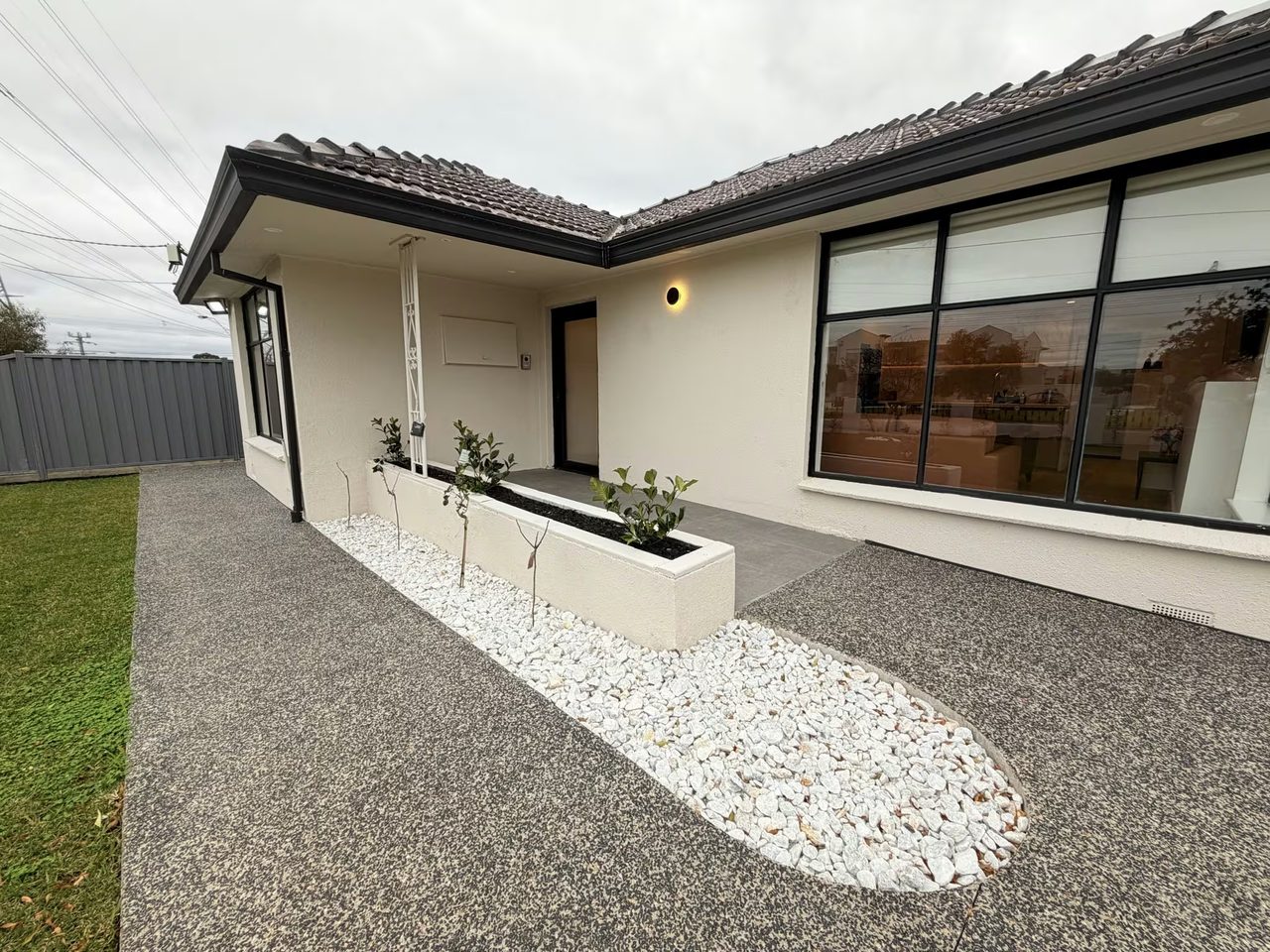 The covered front porch and dark front door, reached by a wide exposed-aggregate path. A rendered planter of young shrubs and a bed of white pebbles run alongside, and a round wall light sits above the door.