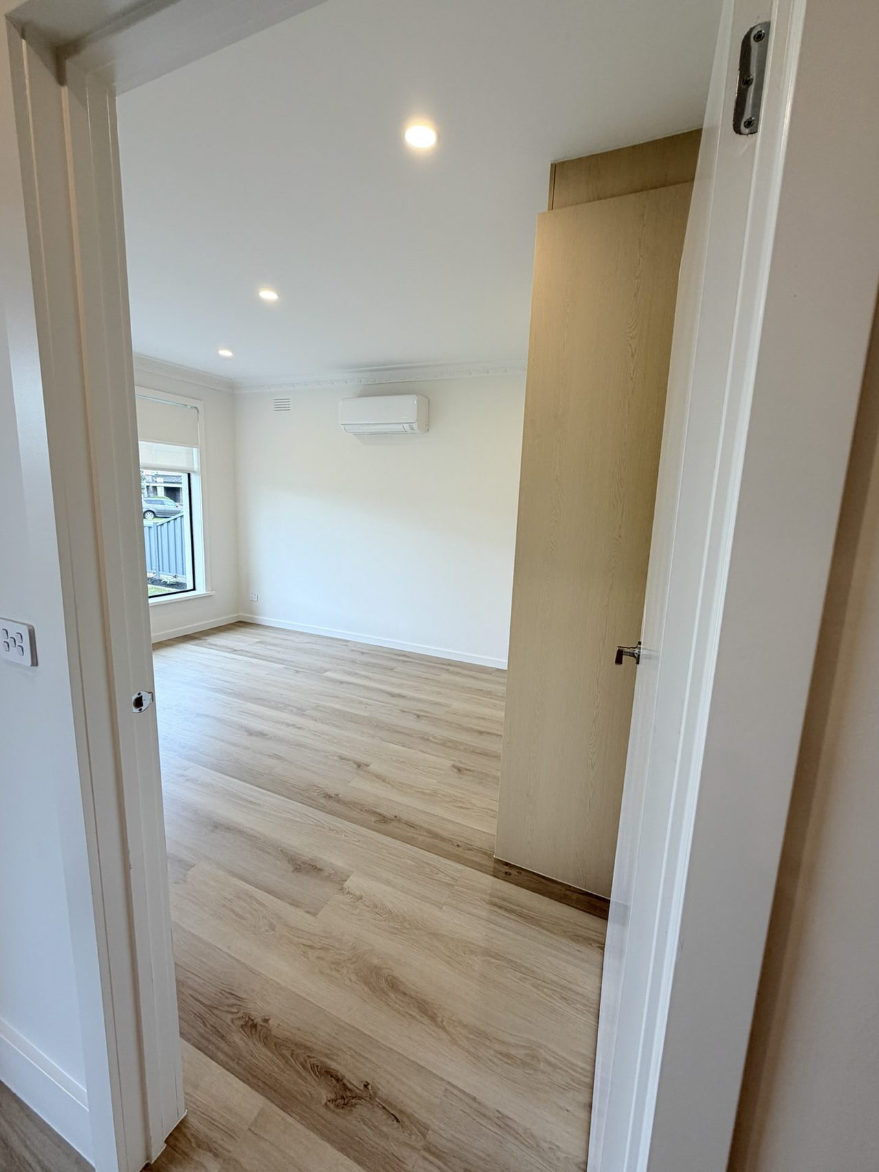 Bedroom one seen from the hall doorway, showing the width of the door opening. Inside are a window with a roller blind, a split-system air conditioner, and unbroken timber-look flooring that continues through the doorway without a threshold strip.