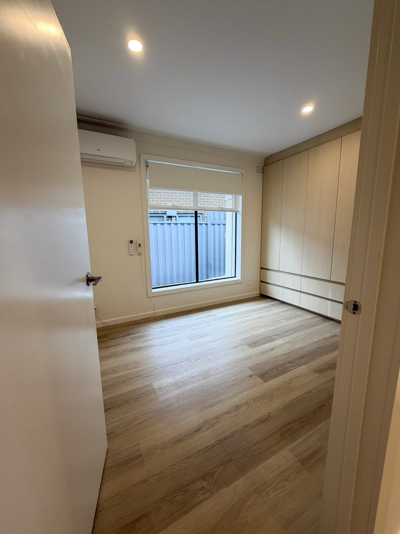 Bedroom two, with a large window and roller blind, a split-system air conditioner, and a built-in wardrobe with drawers along the right-hand wall. The door opens wide onto level vinyl plank flooring.
