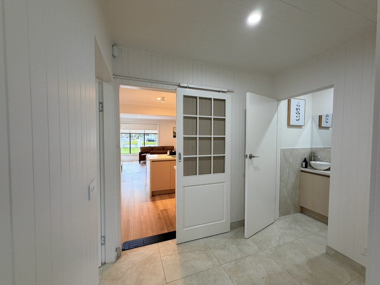 The rear foyer, lined in white vertical panelling, with a sliding barn-style door opening to the living area and a black threshold ramp levelling the small step at that doorway. To the right, the second bathroom stands open, showing a stone-topped vanity with a vessel basin and a wall mirror.