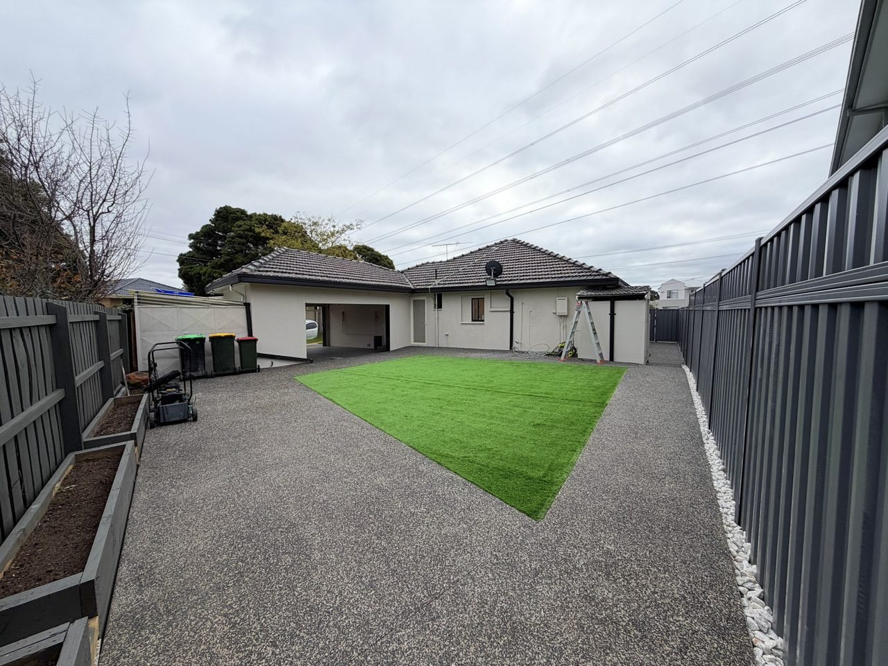 The rear yard, paved end to end in level exposed aggregate with a rectangle of artificial lawn at its centre. A garage and carport sit across the back of the block behind timber and Colorbond fencing, with the paving running flat from the house to both.